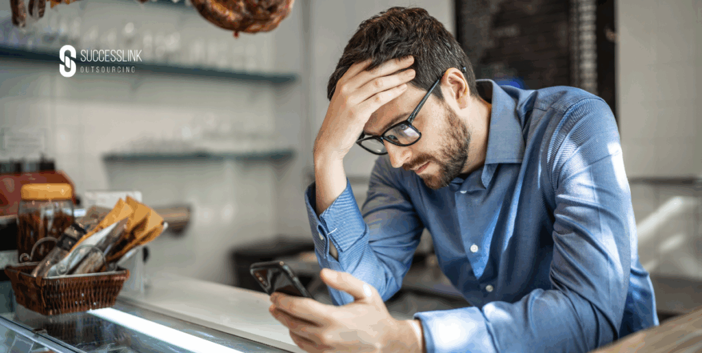 Stressed UK small business owner struggling with admin tasks on smartphone inside café, showing the need for outsourcing support to manage operations effectively.
